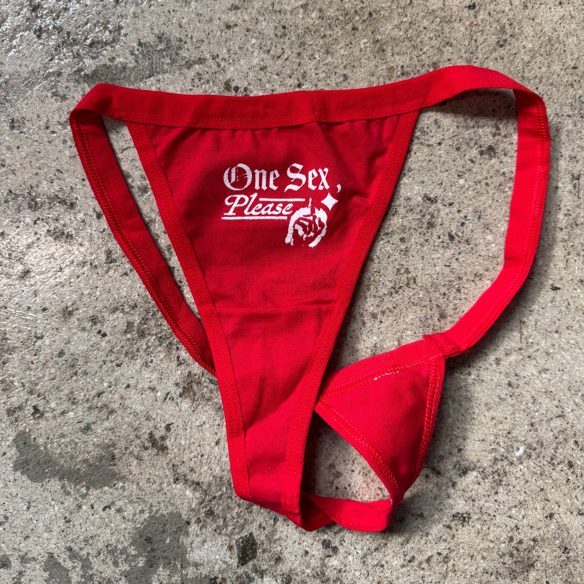Red thong with 'One Sex Please' text on a concrete surface