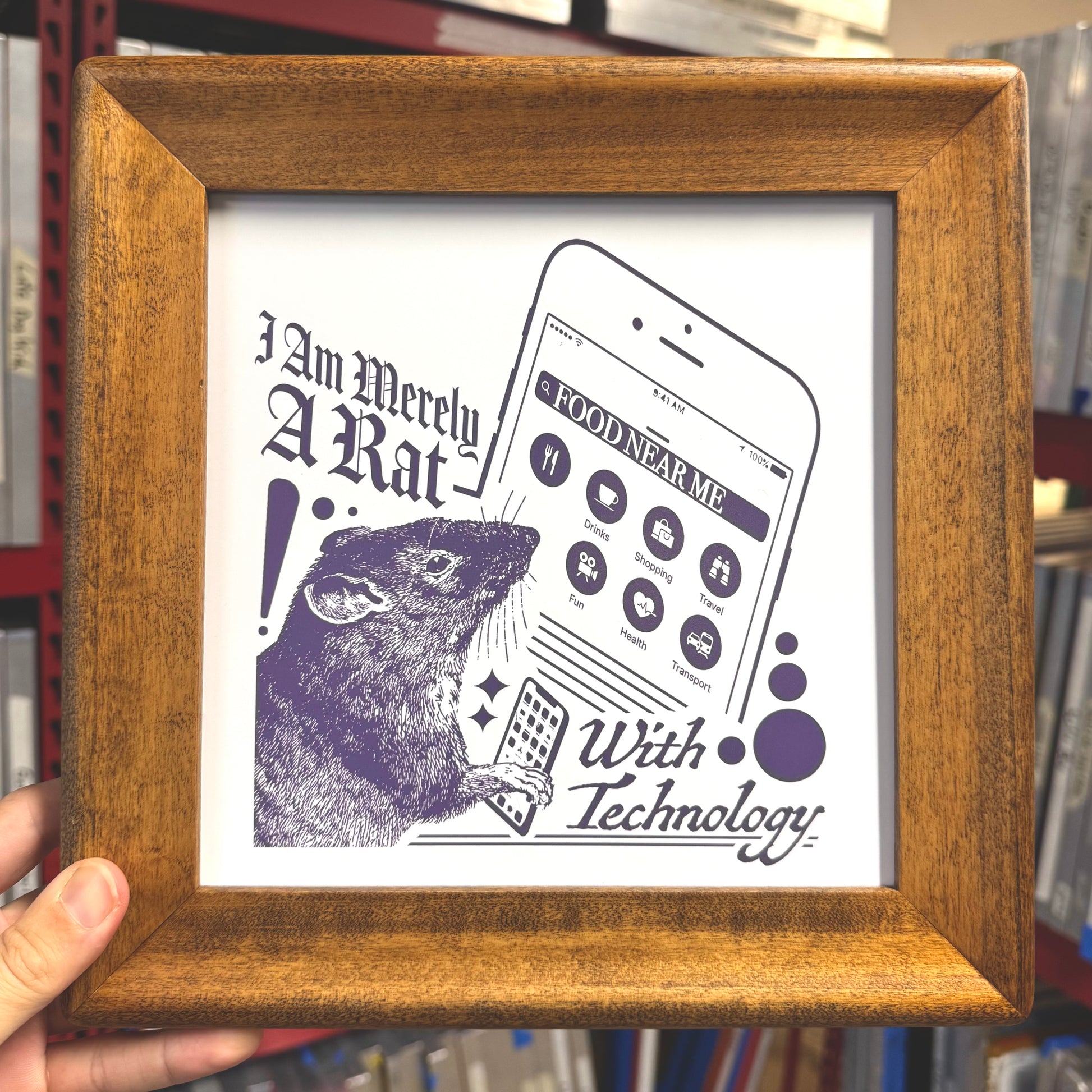 Framed artwork with a rat illustration and smartphone graphic, held in front of bookshelves.