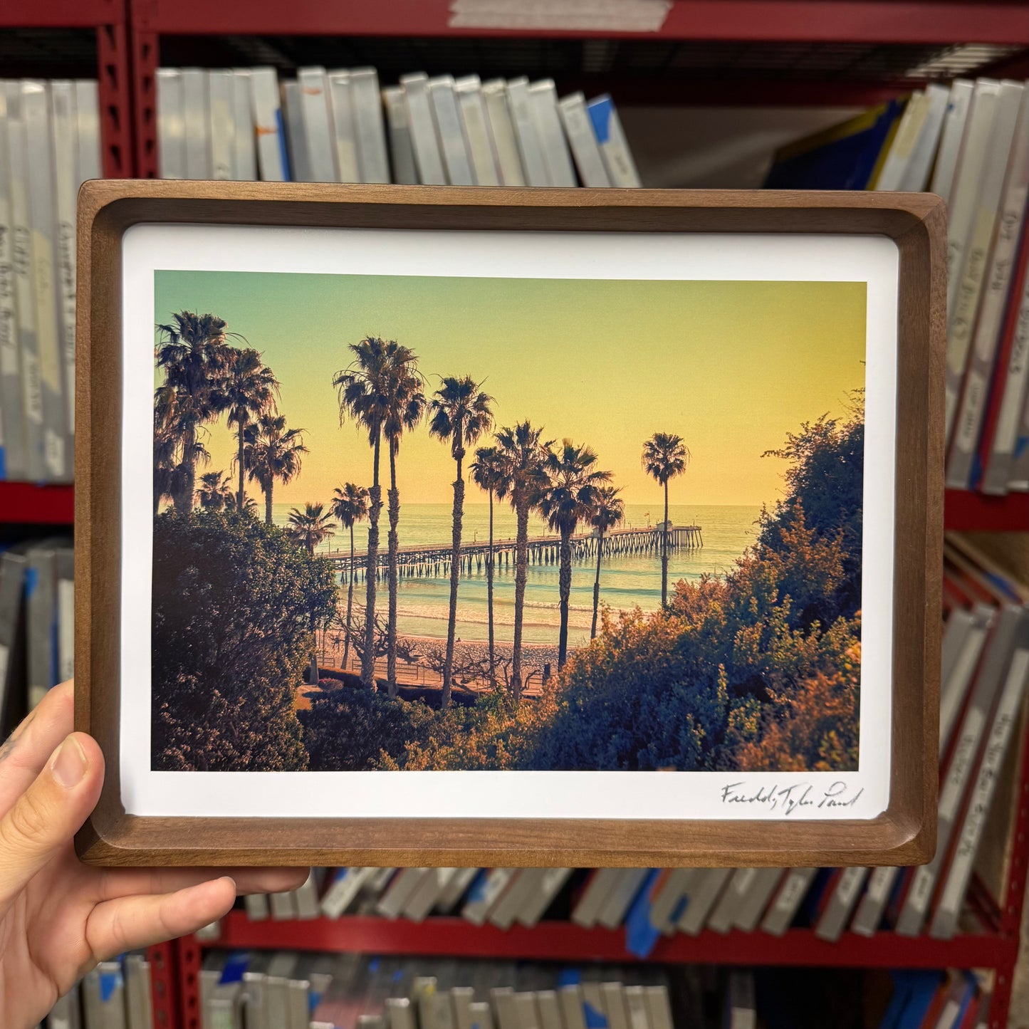 Framed artwork of a beach scene with palm trees and a pier, held in front of a bookshelf.