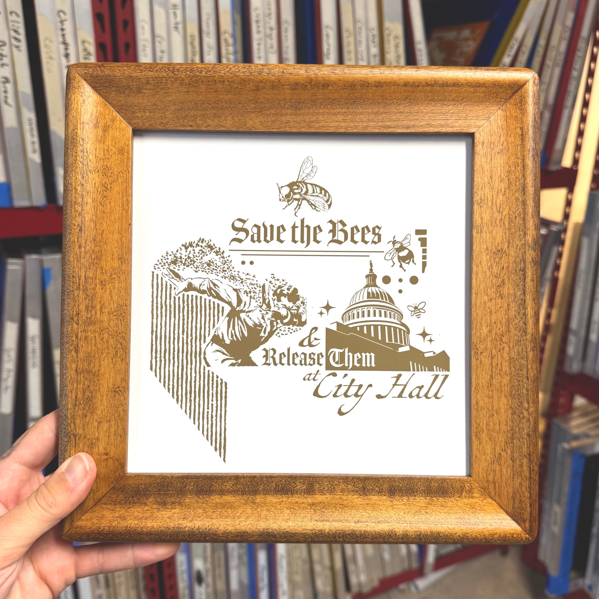 Wooden framed print with 'Save the Bees' text and graphic, held in front of bookshelf.