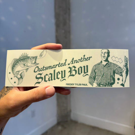 Hand holding a bumper sticker with a graphic of a man and fish, text 'Outsmarted Another Scales Boy' and 'Freddy Tyler Paul'.