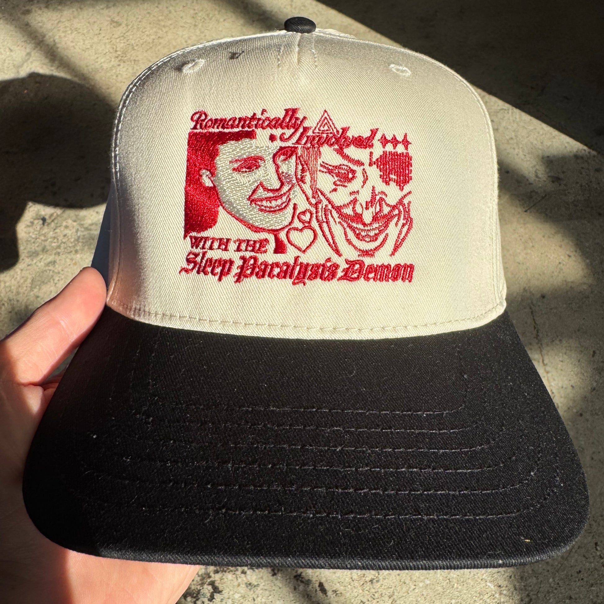 Cap with red graphic and text held by a hand on a concrete surface