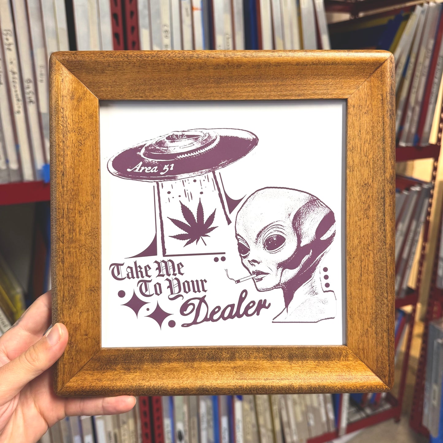 Framed artwork with alien and marijuana leaf design held in front of a bookshelf.