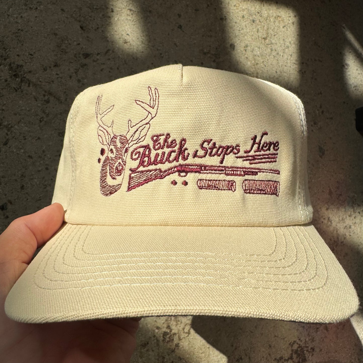 Beige cap with 'The Buck Stops Here' text and deer design held by a hand against a dark background