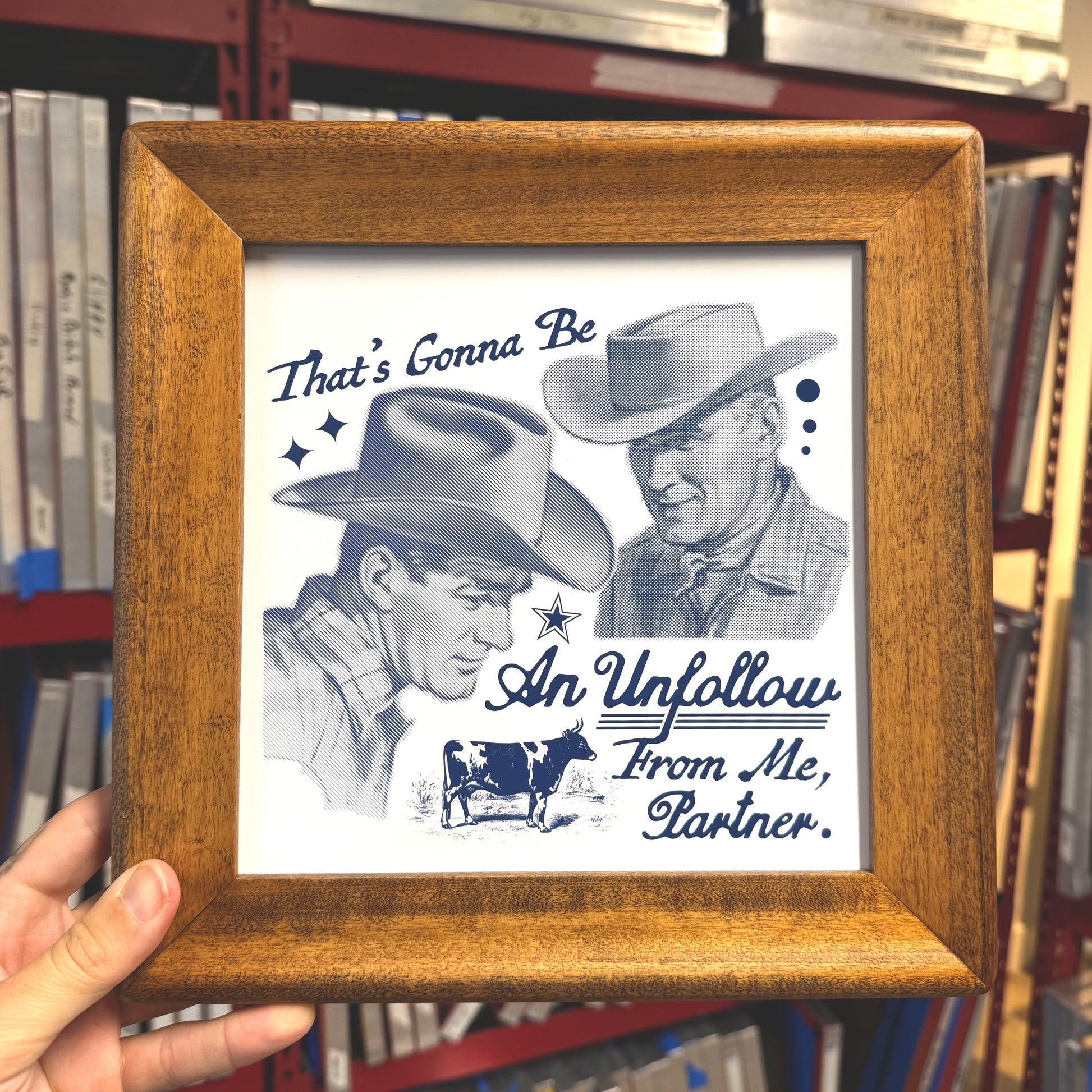 Framed picture with a black and white illustration of two cowboys and text, held in front of a bookshelf.